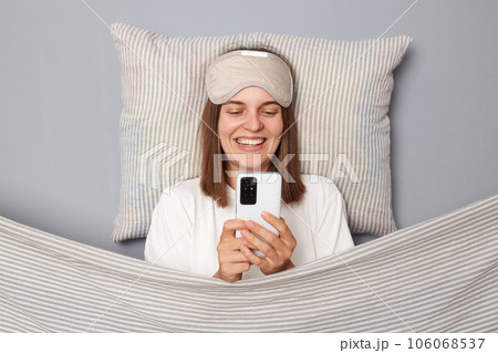 Happy charming woman in white T-shirt and sleeping eye mask lie in bed on pillow under blanket isolated on gray background using cell phone reading internet news smiling happily Happy charming woman in white T-shirt and sleeping eye mask lie in bed on pillow under blanket isolated on gray background using cell phone reading internet news smiling happily 106068537