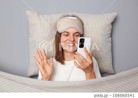 Cute smiling woman in white T-shirt and sleeping eye mask lie in bed on pillow under blanket isolated on gray background having video call waving hand hello wakeup blog. Cute smiling woman in white T-shirt and sleeping eye mask lie in bed on pillow under blanket isolated on gray background having video call waving hand hello wakeup blog. 106068541