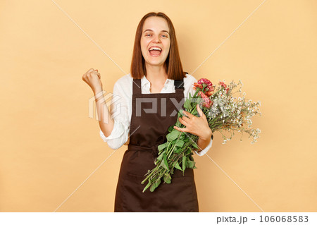 Extremely happy smiling cheerful woman florist wearing brown apron holding bouquet of flowers standing isolated over beige background clenched fists celebrating her business success. Extremely happy smiling cheerful woman florist wearing brown apron holding bouquet of flowers standing isolated over beige background clenched fists celebrating her business success. 106068583