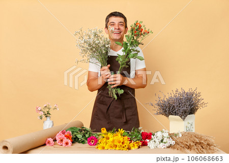 Creative floral arrangements. Beautiful plant collection. Florist's successful market. Cheerful man florist wearing brown apron making bouquet in flower shop standing isolated over beige background Creative floral arrangements. Beautiful plant collection. Florist's successful market. Cheerful man florist wearing brown apron making bouquet in flower shop standing isolated over beige background 106068663