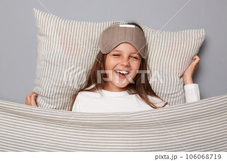 Happy cheerful cute girl wearing sleep eye mask resting on pillow under blanket isolated over gray background smiling while waking up expressing positive emotions 106068719
