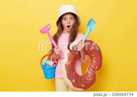 Shocked amazed little girl in panama holding sandbox toys, bucket and rubber ring isolated over yellow background posing with scoop and rake keeps mouth widely opened. Shocked amazed little girl in panama holding sandbox toys, bucket and rubber ring isolated over yellow background posing with scoop and rake keeps mouth widely opened. 106068734