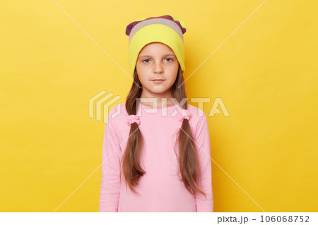 Serious confident little girl with ponytails wearing pink shirt and beanie hat posing isolated over yellow background looking at camera with displeased expression. 106068752