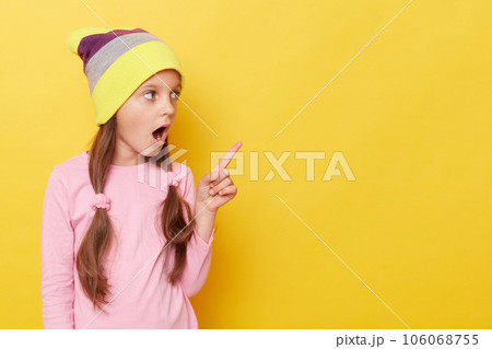 Shocked little girl with ponytails wearing beanie hat standing isolated over yellow background pointing away at advertisemet area being surprised. 106068755