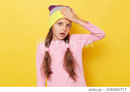 Despair shocked little girl with ponytails wearing pink shirt and beanie hat standing isolated over yellow background having troubles showing facepalm gesture. 106068757