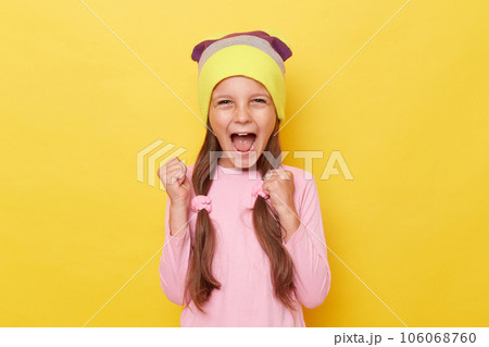 Extremely happy cheerful little girl with ponytails wearing pink shirt and beanie hat posing isolated over yellow background clenched fist rejoicing her success. Extremely happy cheerful little girl with ponytails wearing pink shirt and beanie hat posing isolated over yellow background clenched fist rejoicing her success. 106068760