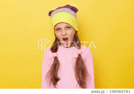 Deleterious little girl with ponytails wearing pink shirt and beanie hat standing isolated over yellow background screaming expressing negative emotions. 106068761