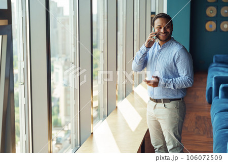 Smiling businessman is talking phone with client while standing in modern coworking near window Smiling businessman is talking phone with client while standing in modern coworking near window 106072509