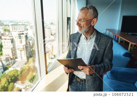 Pensive senior entrepreneur holding clipboard and looking at window while standing in coworking Pensive senior entrepreneur holding clipboard and looking at window while standing in coworking 106072521