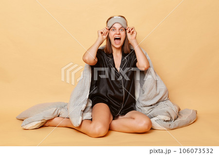 Excited amazed woman sitting on floor wrapped in plaid wearing pajama, sleep eye mask isolated over beige background taking off blindfold with excitement. Excited amazed woman sitting on floor wrapped in plaid wearing pajama, sleep eye mask isolated over beige background taking off blindfold with excitement. 106073532