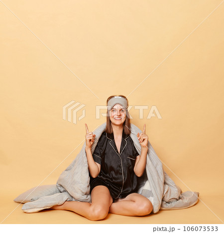 Cheerful joyful woman sitting on floor wrapped in plaid wearing pajama, sleep eye mask isolated over beige background pointing up at copy space for advertisement. Cheerful joyful woman sitting on floor wrapped in plaid wearing pajama, sleep eye mask isolated over beige background pointing up at copy space for advertisement. 106073533