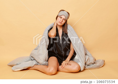 Tired exhausted woman sitting on floor wrapped in plaid wearing pajama, sleep eye mask isolated over beige background feeling pain in neck after uncomfortable sleep. Tired exhausted woman sitting on floor wrapped in plaid wearing pajama, sleep eye mask isolated over beige background feeling pain in neck after uncomfortable sleep. 106073534