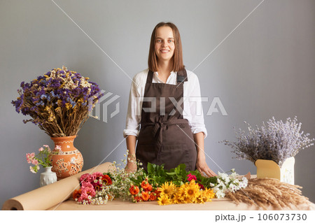 Professional florist workshop. Fresh flower bouquet design. Fragrant organic arrangements. Smiling confident woman florist at workplace in flower shop standing isolated over gray background Professional florist workshop. Fresh flower bouquet design. Fragrant organic arrangements. Smiling confident woman florist at workplace in flower shop standing isolated over gray background 106073730
