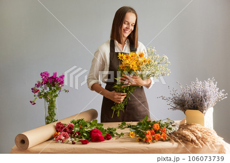 Skilled floral craftsmanship. Delicate flower compositions. Hard working woman florist creating beautiful bouquet at workplace in flower shop standing isolated over gray background Skilled floral craftsmanship. Delicate flower compositions. Hard working woman florist creating beautiful bouquet at workplace in flower shop standing isolated over gray background 106073739