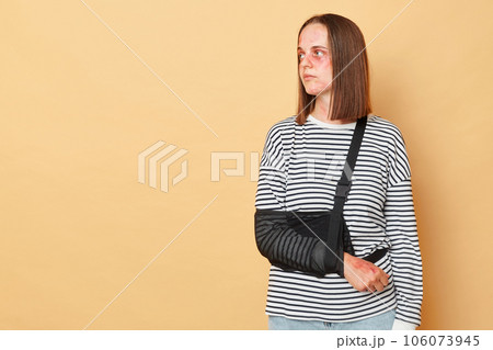 Unhappy disabled woman with bruises and injuries wearing striped shirt standing isolated over beige background looking away at empty space for promotion. 106073945