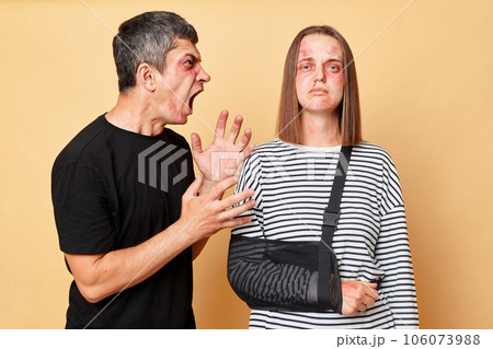 Home violence. Relationship arguingg. Aggressive man screaming with hate and anger, beating his woman, wife with bruise and bandage isolated over beige background. Home violence. Relationship arguingg. Aggressive man screaming with hate and anger, beating his woman, wife with bruise and bandage isolated over beige background. 106073988