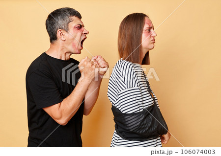 Aggressive man shouting with anger abusing his wife, battered woman with bruise turning way from husband with sad face isolated over beige background 106074003