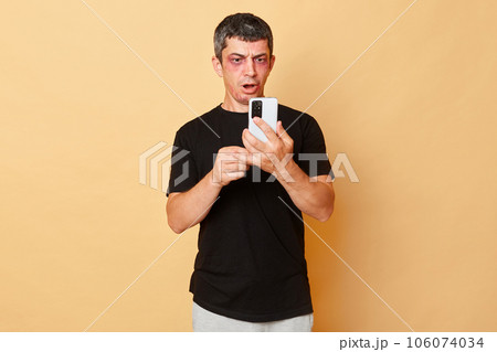 Traumatized beaten man in black T-shirt with bruises and abrasions on his face isolated over beige background using mobile phine chatting with psychologist calling police. Traumatized beaten man in black T-shirt with bruises and abrasions on his face isolated over beige background using mobile phine chatting with psychologist calling police. 106074034