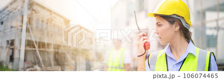Young attractive construction woman in safety vest with yellow helmet working with radio, standing on building construction site. Home building project. 106074465