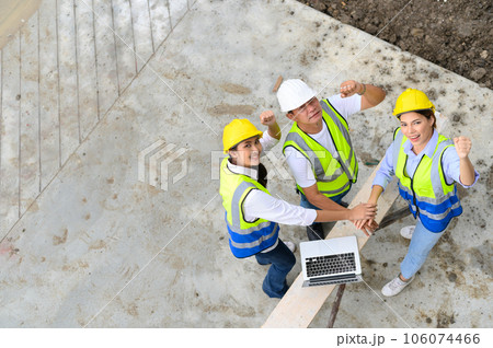 Top view of contractors, engineers and formats team in safety vests with helmets working with laptops, standing on under-construction building site. Home building project. look at the camera Top view of contractors, engineers and formats team in safety vests with helmets working with laptops, standing on under-construction building site. Home building project. look at the camera 106074466