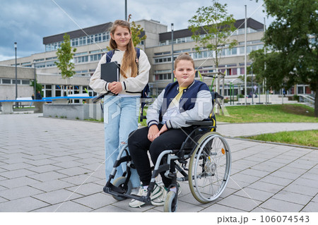 Portrait of classmates, boy in wheelchair and girl with backpack, outdoor near school building 106074543