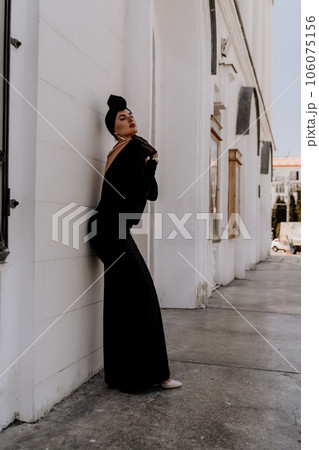Stylish woman in the city. Fashion photo of a beautiful model in an elegant black dress posing against the backdrop of a building on a city street Stylish woman in the city. Fashion photo of a beautiful model in an elegant black dress posing against the backdrop of a building on a city street 106075156