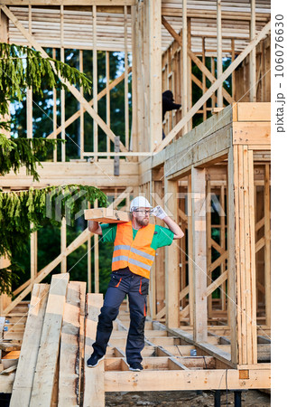 Carpenter building wooden-framed house near the forest. Man holding a large beam on his shoulder, dressed in work clothes and helmet. Idea of modern and sustainable construction. 106076630
