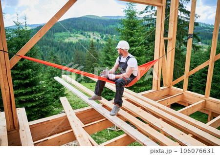 Carpenter, working on construction of wooden-framed house, takes break in hammock, dressed in protective helmet, work overalls and gloves. He enjoys picturesque view of mountains and forests. Carpenter, working on construction of wooden-framed house, takes break in hammock, dressed in protective helmet, work overalls and gloves. He enjoys picturesque view of mountains and forests. 106076631