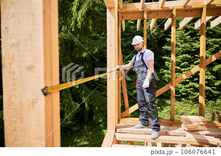 Carpenter constructing wooden skeleton building. Man measures distances with tape measure while wearing work attire and helmet. The concept of modern, environmentally-friendly construction. 106076641