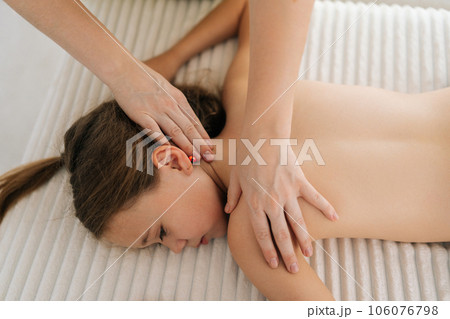Top view of cute little girl having neck, shoulder and back massage by unrecognizable female masseuse lying on massage table. Adorable preteen kid getting physiotherapy from masseuse for body care. Top view of cute little girl having neck, shoulder and back massage by unrecognizable female masseuse lying on massage table. Adorable preteen kid getting physiotherapy from masseuse for body care. 106076798