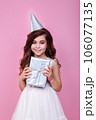 Smiling kid girl in birthday party princess hat holding gift box on a pink background 106077135