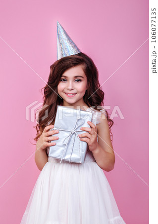 Smiling kid girl in birthday party princess hat holding gift box on a pink background Smiling kid girl in birthday party princess hat holding gift box on a pink background 106077135