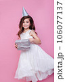 Smiling kid girl in birthday party princess hat holding gift box on a pink background. Kid birthday 106077137
