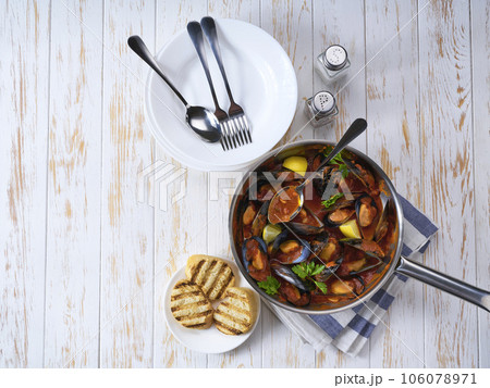 Steamed mussels in shells and tomato sauce with roasted bread and greens on a white table. Seafood dinner. 106078971