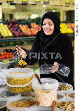 Asian girl in hijab buys pickled olives by weight in supermarket Asian girl in hijab buys pickled olives by weight in supermarket 106080400
