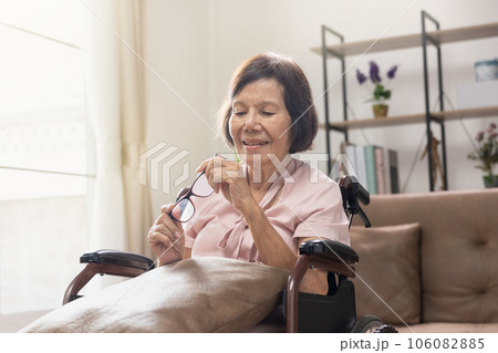 Senior woman holding eyeglasses sitting on wheelchair at home 106082885