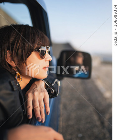 夕焼けの海岸沿い運転席に座ったサングラスの女性がバックミラーに手を伸ばしてる 夕焼けの海岸沿い運転席に座ったサングラスの女性がバックミラーに手を伸ばしてる 106089384