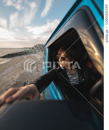 夕焼けの海岸沿い運転席に座ったサングラスの女性がバックミラーに手を伸ばしてる 106089389