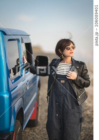 オーバーオールにライダースサングラス姿の女性が車の横で遠くを見ながら立っている 106089401