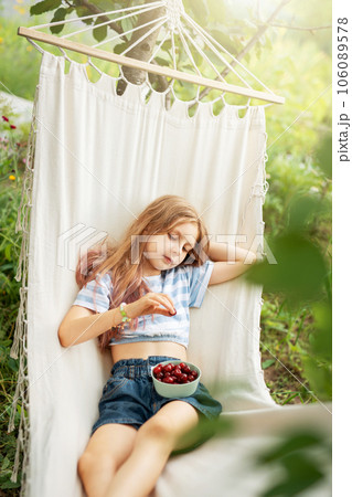 A little girl rests in a hammock and eats cherries in the summer. 106089578