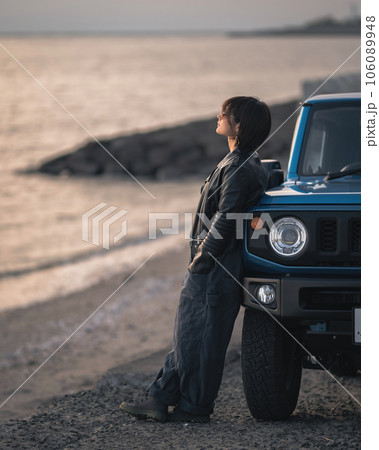 海に沈む夕日を見ながらオーバーオールにライダース姿の女性が車にもたれて黄昏てる 106089948