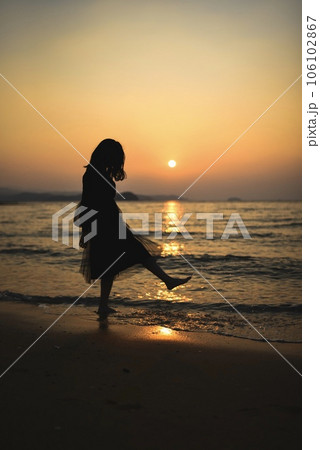 夕日をバックに海岸で女性が水を蹴り上げてるシルエットの写真素材