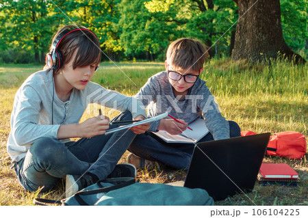 Boy in glasses carefully watching pupil in earphones who reading task from notebook, classmates study in park together. 106104225