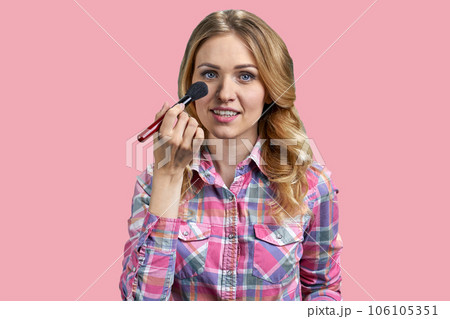 Attractive young woman applying make-up brush to her face. Isolated on pink. Attractive young woman applying make-up brush to her face. Isolated on pink. 106105351