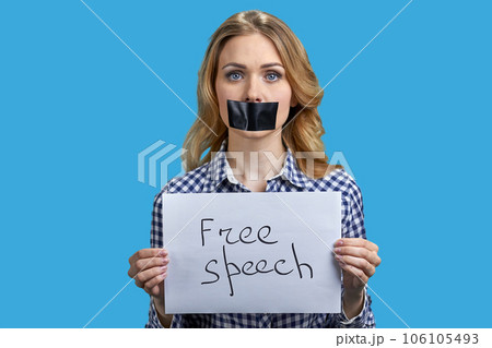 Young blonde woman protester with taped mouth holding banner with inscription free speech. Silenced woman on blue background. Young blonde woman protester with taped mouth holding banner with inscription free speech. Silenced woman on blue background. 106105493