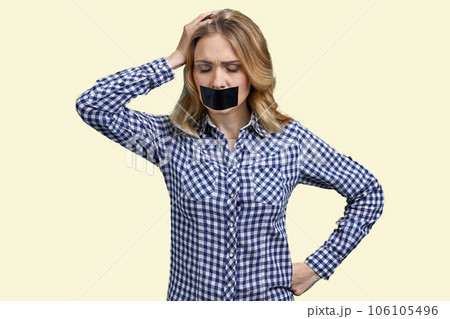 Portrait of young desperate blond woman with her mouth taped. Young lady touchine her head. Isolated on pastel yellow background. 106105496