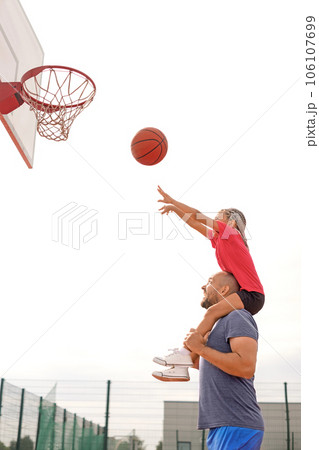 A child throws a ball into a basketball hoop, sitting on his father's shoulders. Dad teaches the child to play basketball. Outdoor sports ground 106107699