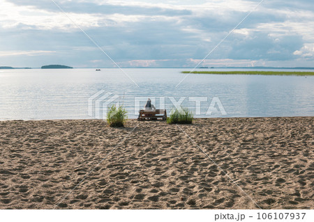 sandy beach on the shore of a vast lake, a woman traveler is sitting on a bench made of old pallets sandy beach on the shore of a vast lake, a woman traveler is sitting on a bench made of old pallets 106107937