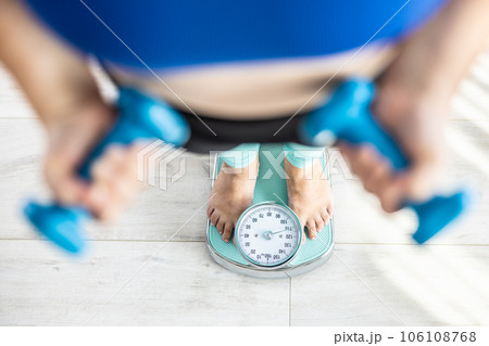 Heavy fat woman stands on a scale and holds dumbbells in her hands Heavy fat woman stands on a scale and holds dumbbells in her hands 106108768