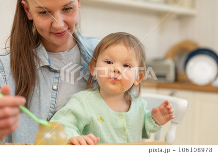 Happy family at home. Mother feeding her baby girl from spoon in kitchen. Little toddler child with messy funny face eats healthy food at home. Young woman mom giving food to kid daughter 106108978